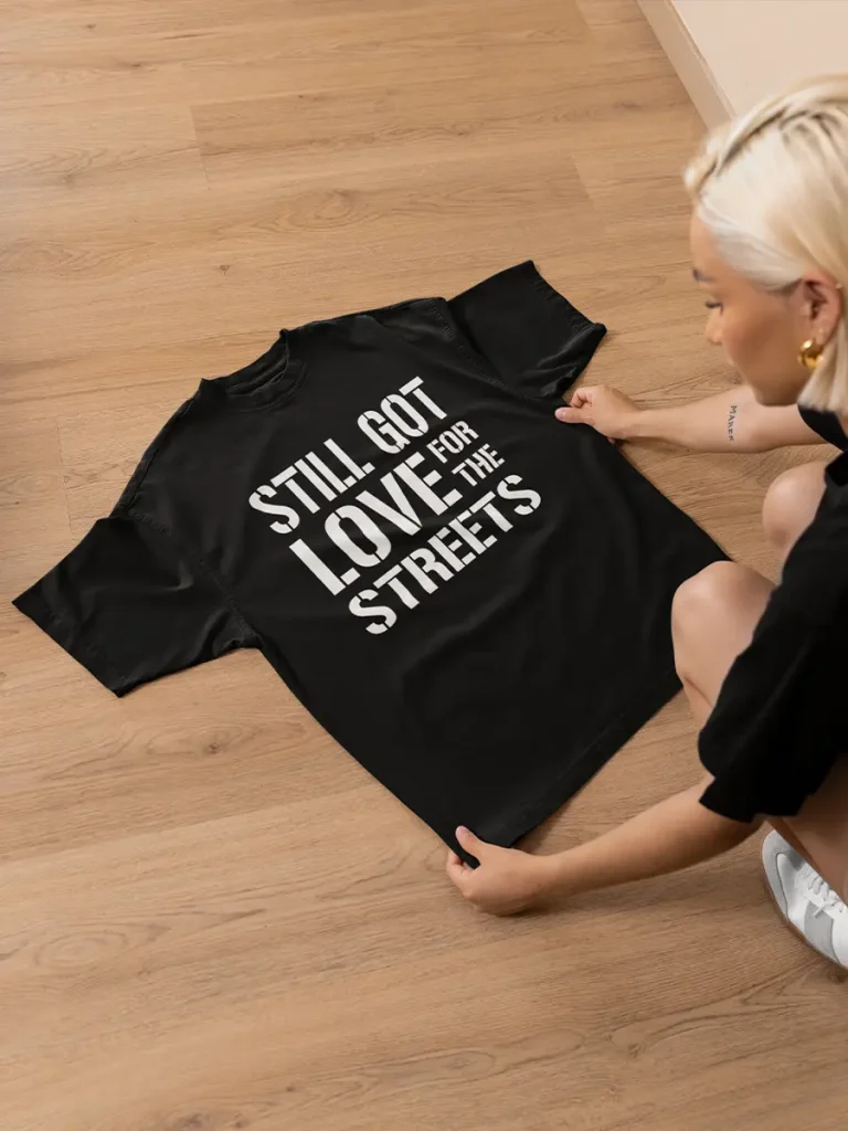A woman holding a black t-shirt with a rap lyric printed on it, which reads "Still Got Love for the Streets"