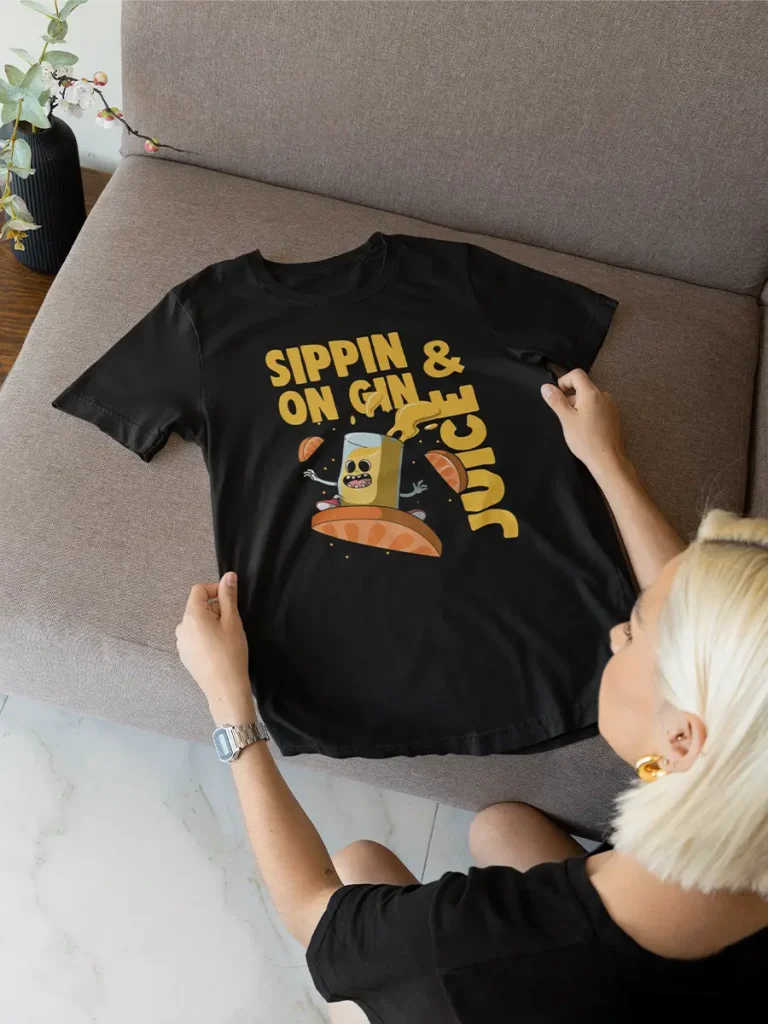 A flat lay of a t-shirt that reads “Sippin on Gin and Juice” stylized in yellow bright font with a juice illustration